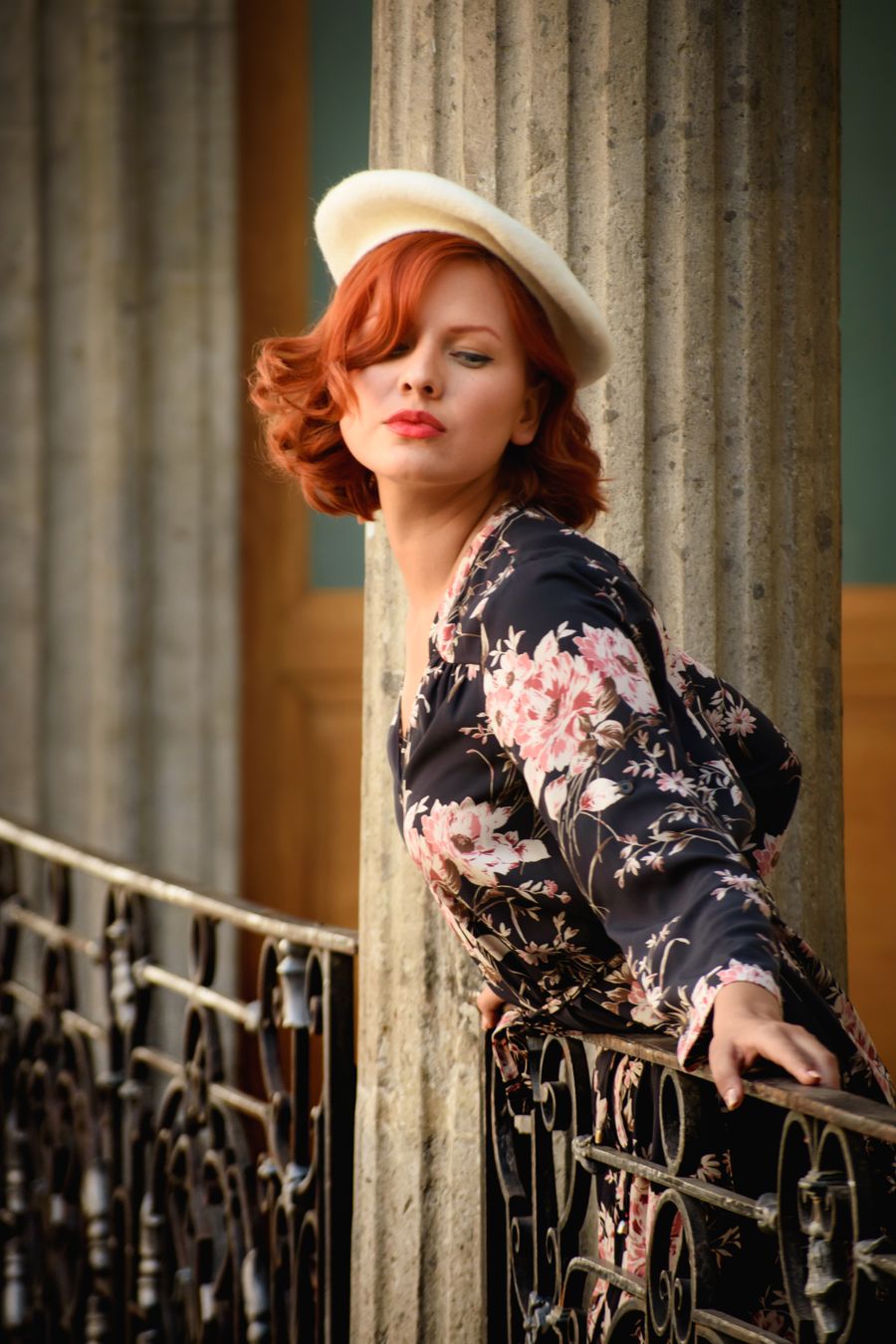 Mujer pelirroja con sombrero claro y vestido oscuro de estampado floral, apoyada en una barandilla de hierro junto a columnas clásicas